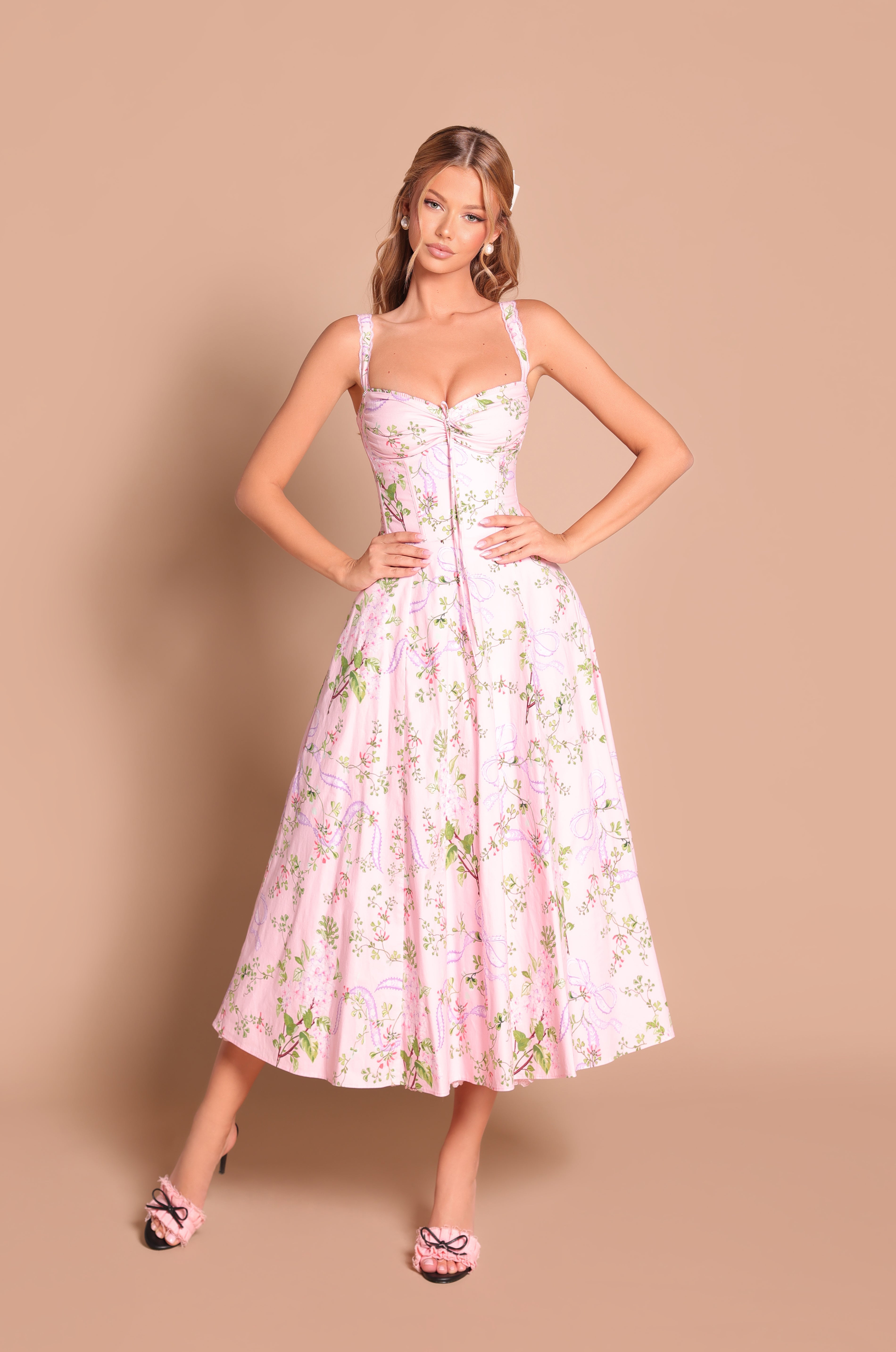 A woman stands against a beige background wearing the Celenia Blush Pink Floral Casual Midi Dress, a fit-and-flare midi with spaghetti straps and a subtle corset bodice, paired with matching pink heeled sandals.